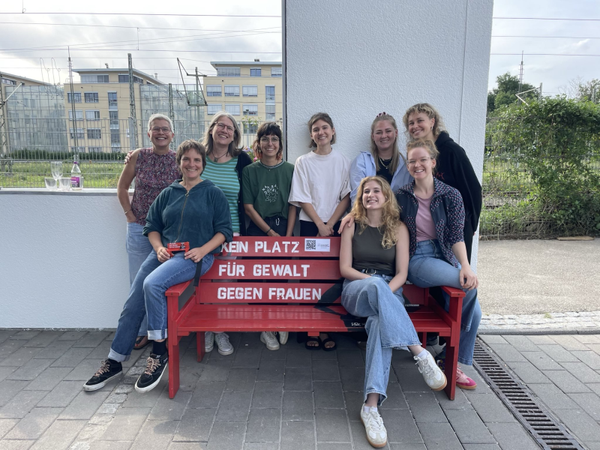 Quelle: HILA DW Freiburg Eine Frauengruppe hat sich um eine rote Sitzbank versammelt